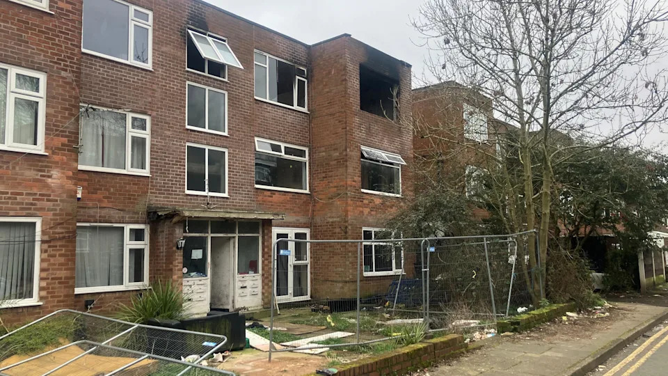 Photograph of an abandoned apartment block on Baguley Crescent. The image shows a damaged fence, a burnt out window and an open front door.