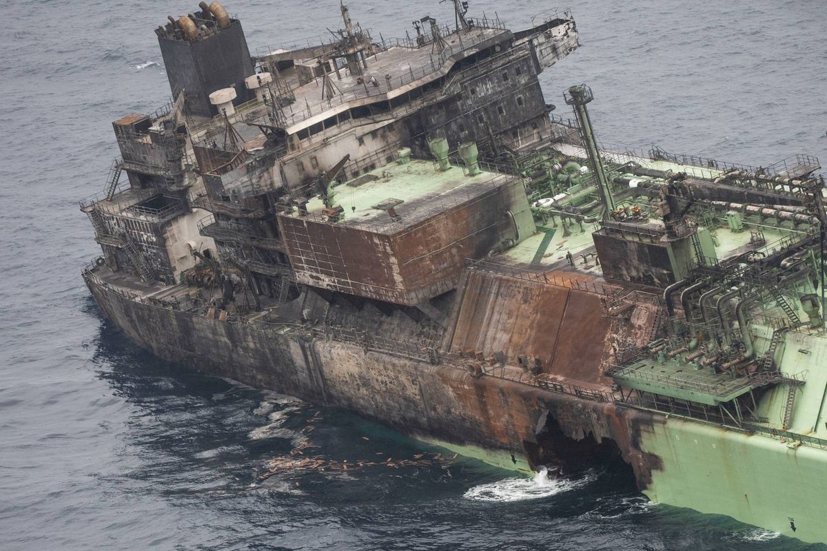 An aerial view depicts a large, deteriorated vessel afloat in the ocean, with visible signs of damage and corrosion, accompanied by a smaller boat nearby.