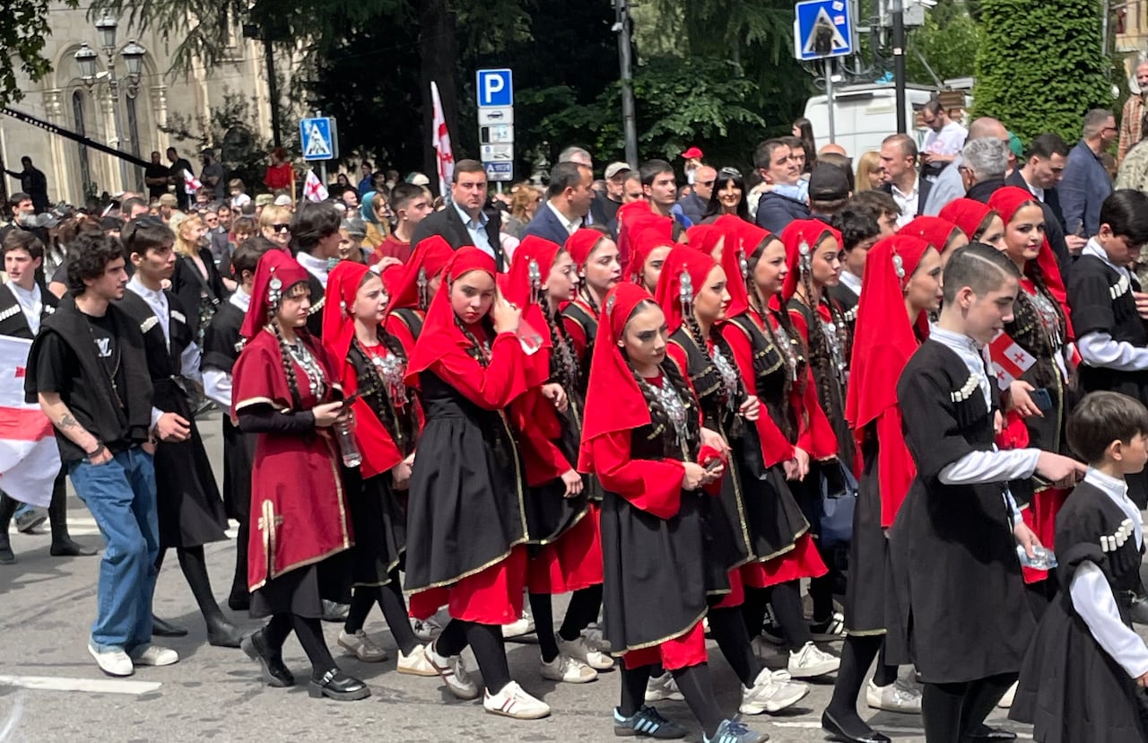 A parade is walking down the street ... young children are walking wearing red and black cloak-like outfits