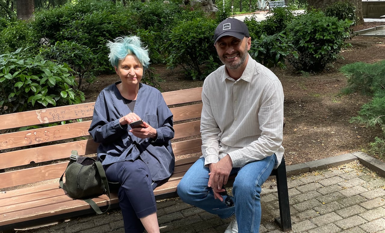 A man and woman are sitting on a bench talking (she is holding a microphone)