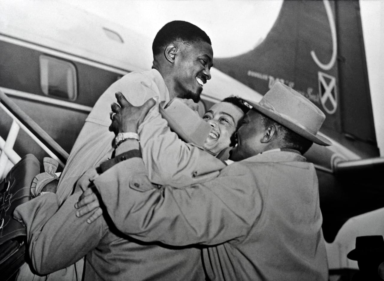 Black and white photo of a smiling man being embraced by a man and woman and he descends from an airplane