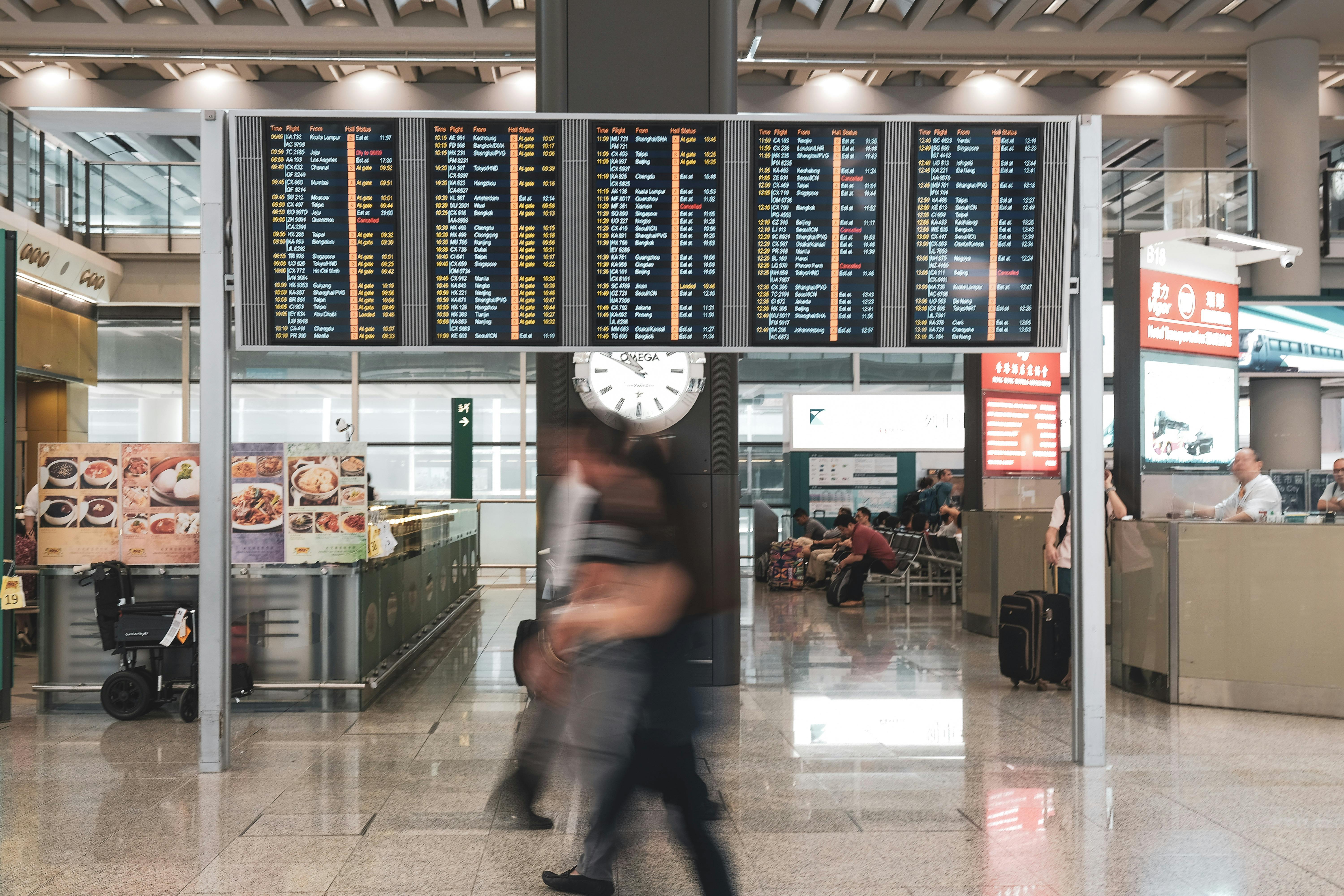 Airport terminal with a large departure board, clock, and passengers moving and waiting with luggage.