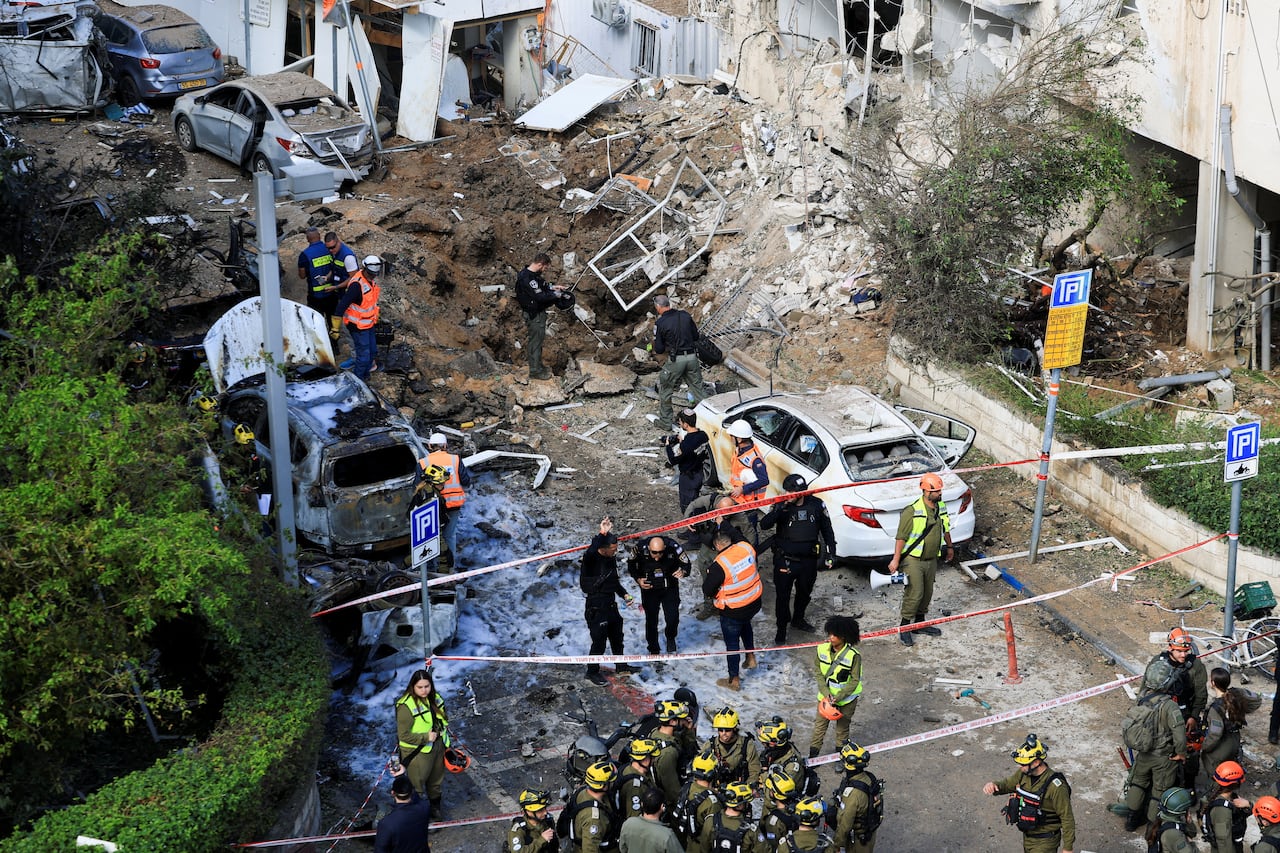 Emergency crews work at a building that is damaged by a missile strike. Burned cars line the street outside the damaged building.