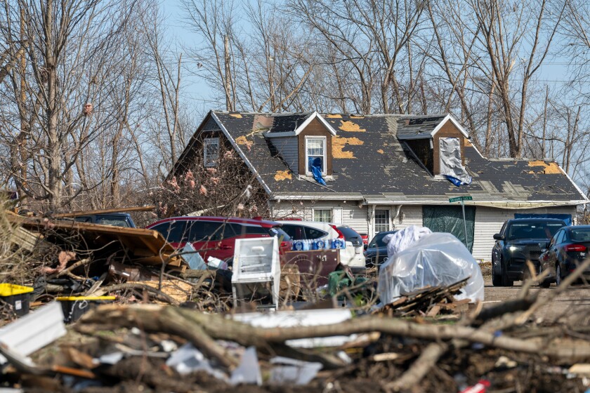 A tornado-damaged home seen from the ground. Downed tree limbs are in the foreground with a damaged home in the back.