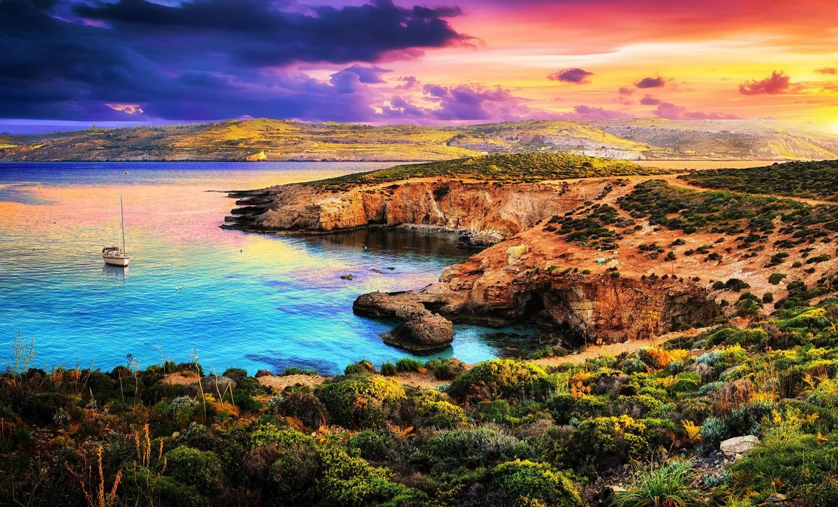 Dramatic sunset over a tranquil bay with a sailboat and rugged coastline,Malta,Comino