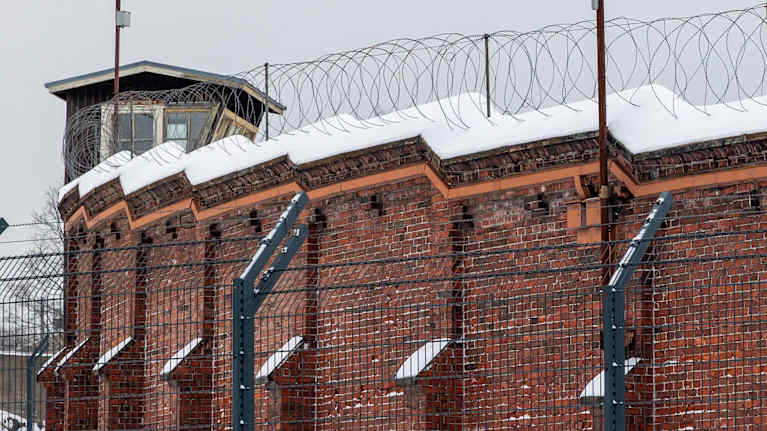 A red-brick wall with snow and barbed wire on top and a guard tower behind it.
