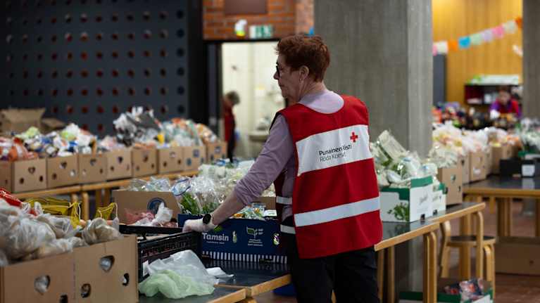 A Finnish Red Cross worker arranging boxes of food laid out on many tables in a large room.