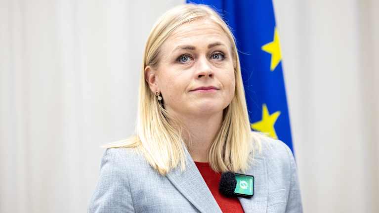 Woman with blonde hair and grey jacket looking up, with an EU flag behind her.