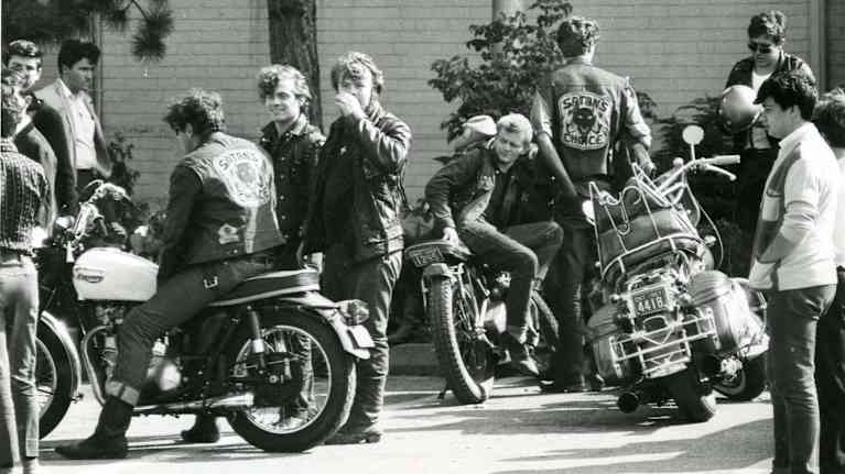 Black and white photo of about a dozen men standing by or sitting on motorcycles on a city street, some with Satan's Choice patches on their jackets.