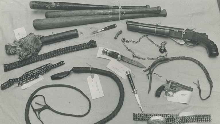 Black and white photo of confiscated weapons including  whips, guns, knives, bicycle chains and baseball bats.