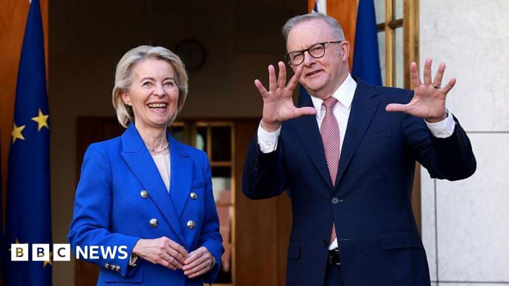 A woman in a bright blue suit smiles next to a man in a dark blue suit who is gesturing with his hands.