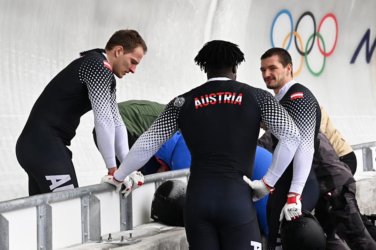 Austria's Daiyehan Nichols-Bardi (C), Austria's Daniel Bertschler (L) and Austria's Sebastian Mitterer (R) react as medics bring off Austria's Jakob Mandlbauer after a crash in the bobsleigh men's 4-man heat 2 at Cortina Sliding Centre during the Milano Cortina 2026 Winter Olympic Games in Cortina d'Ampezzo on February 21, 2026. (Photo by Tiziana FABI / AFP via Getty Images)
