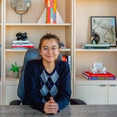 Reporter Joey Hadden in an argyle sweater sits at a desk with light, wooden bookshelves behind her.