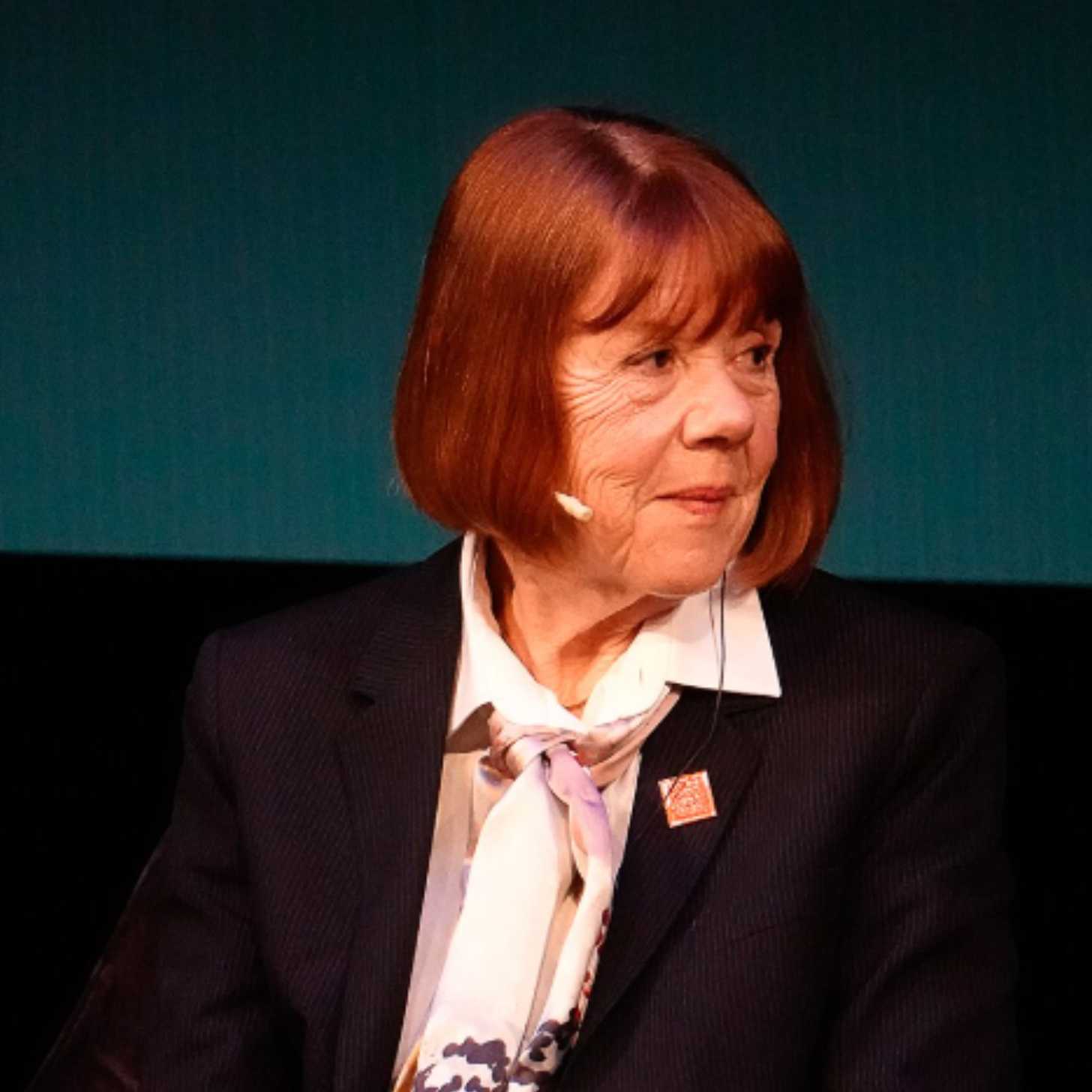 Gisèle Pelicot, an older White woman with red hair, sits on stage for an interview in front of a blue background.