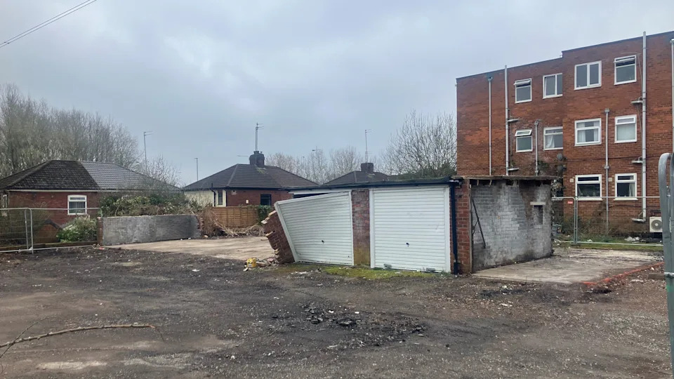 Photograph of abandoned garages on Baguley Crescent in Prestwich. The image shows waste ground in the foreground and an empty block of flats in the background.