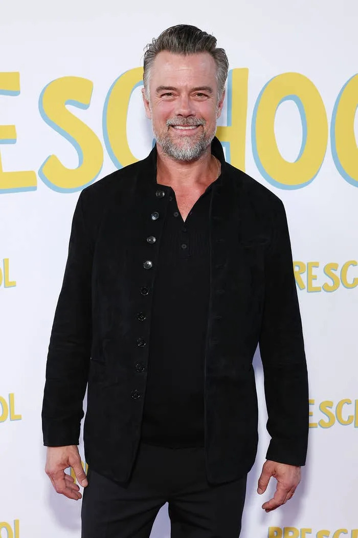 Josh Duhamel on red carpet in black button-up jacket over black top, smiling at a public event