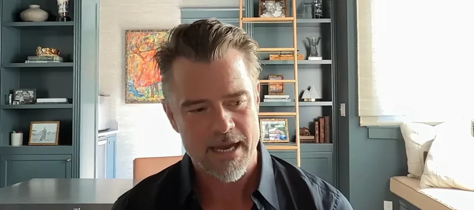 Man with facial hair and styled hair in a home office setting with bookshelves and art in the background
