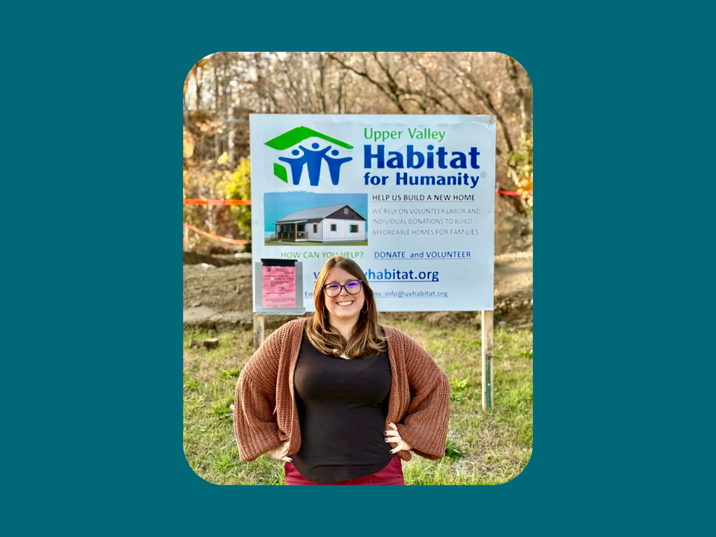 A woman stands smiling in front of an Upper Valley Habitat for Humanity sign at an outdoor construction site.