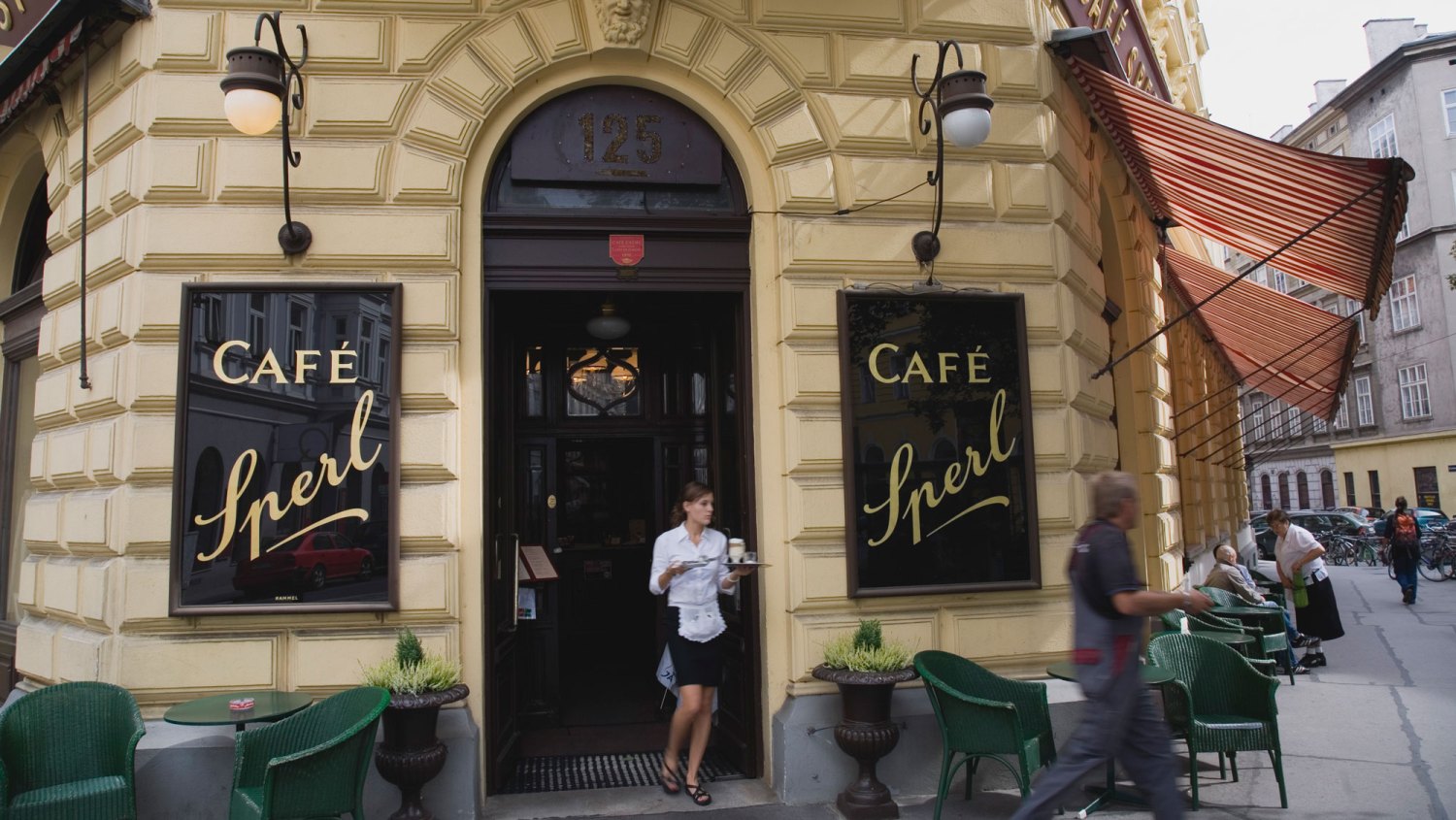 Mariahilf District Cafe Sperl Exterior facade with waitress carrying coffee. Austria, Vienna, Mariahilf District. Cafe Sperl the preferred cafe of Adolf Hitler. Exterior facade with waitress carrying coffee order at entrance. Striped awning pulled out at side with customers sitting at outside tables on street below.