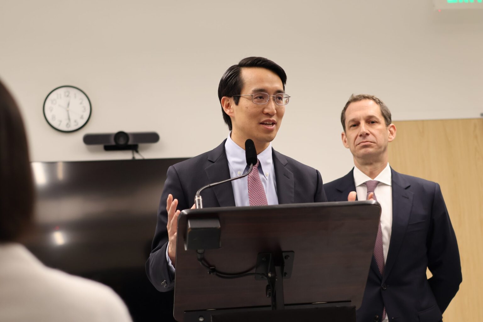 A person speaking at a podium with a microphone, wearing a suit and tie. Another person stands behind, also in a suit. A wall clock and TV are in the background.