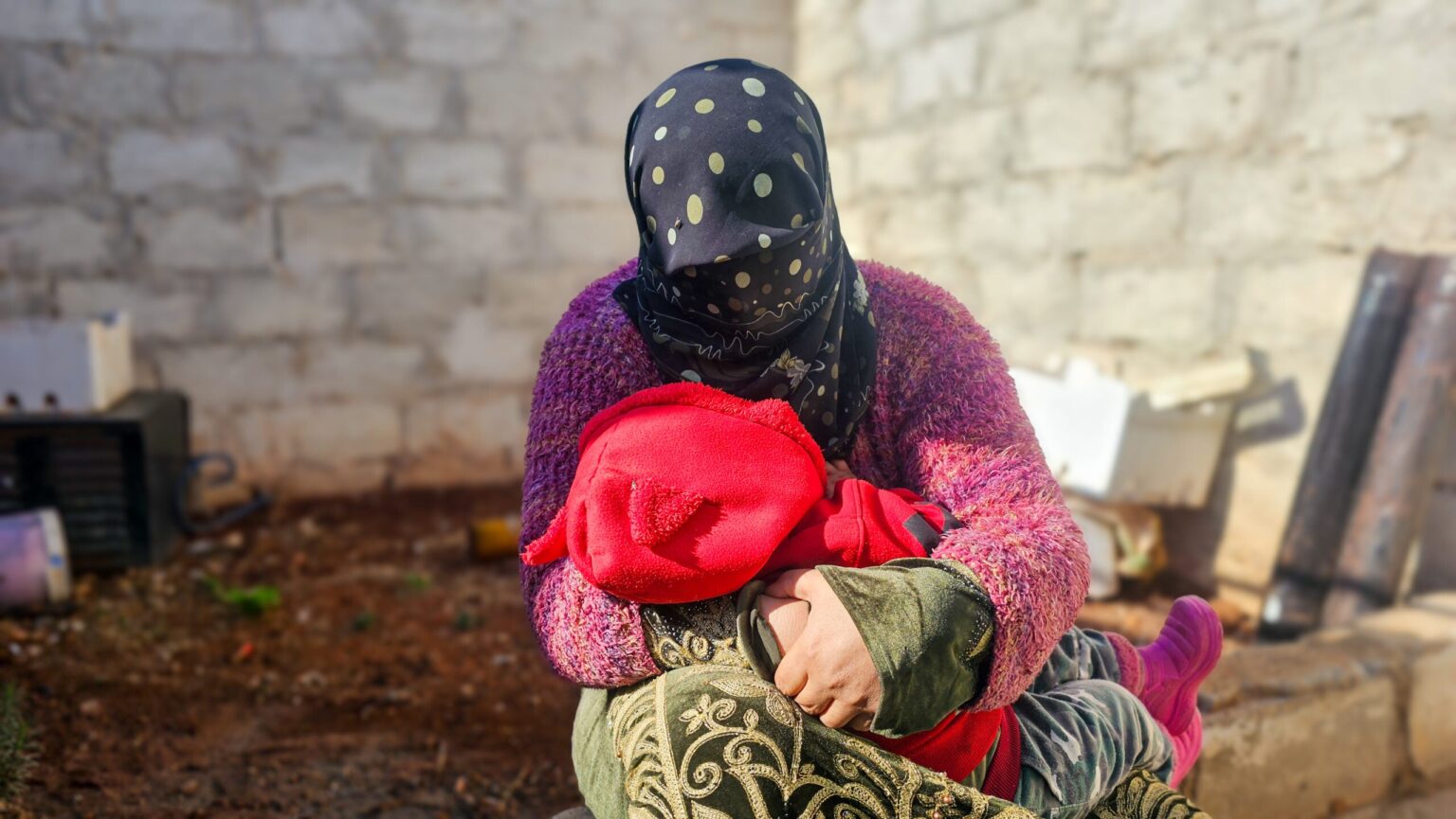 In Syria, women and children carry the burden when funding is cut A woman in a dark headscarf cradles a small child in a read sweatshirt.