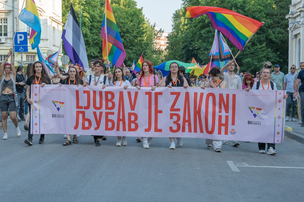 People waving rainbow flags and walking behind a banner at a Pride parade in Sarajevo.