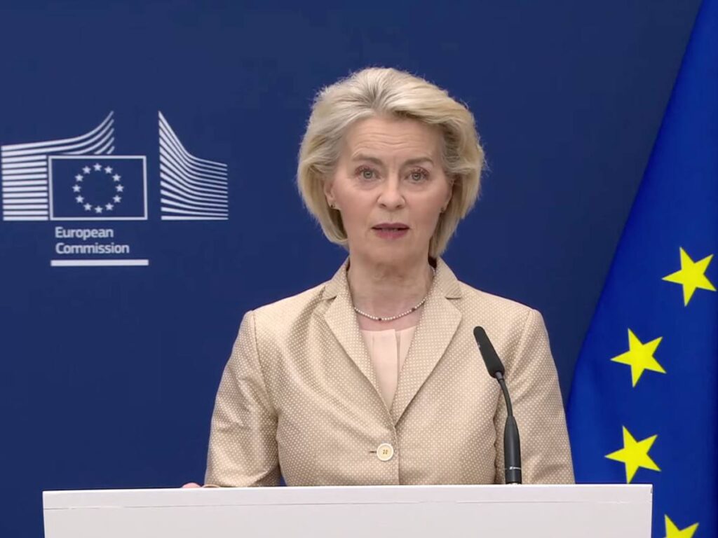 blonde woman (Ursula von der Leyen) stands at a dais with EU branding behind