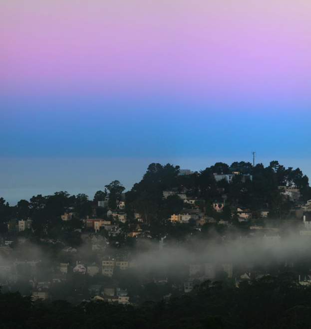 Layers of purple and blue and gray are seen in a clear sky over a hilly city full of trees