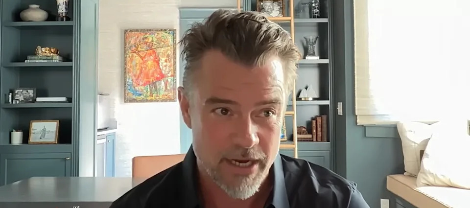 A person with short hair and a beard speaks in a modern room with shelves and a colorful painting in the background