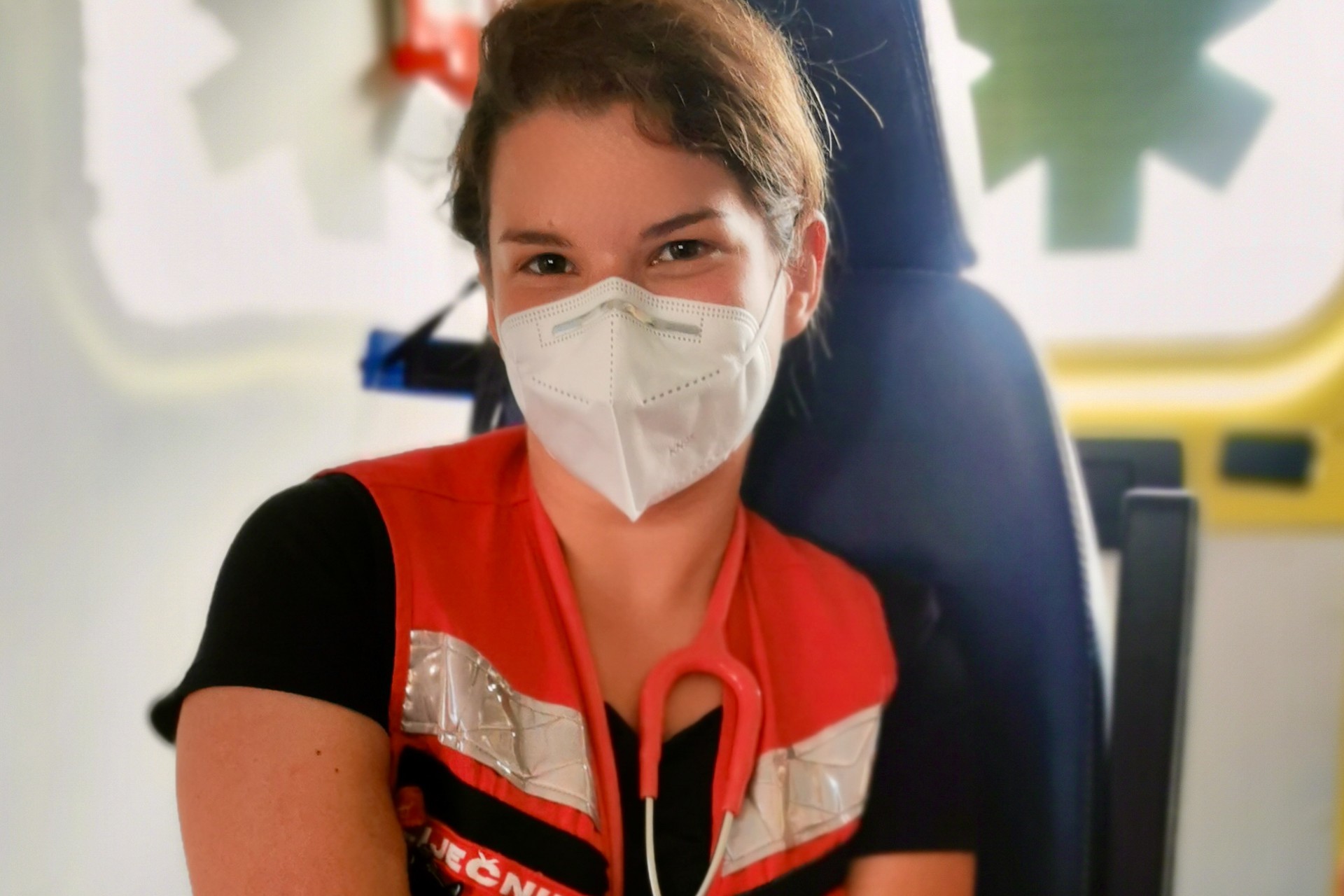 A healthcare worker wearing a face mask and red vest sitting inside an ambulance