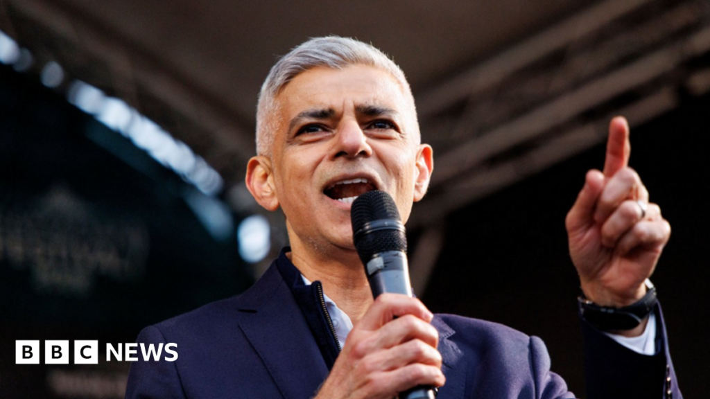 Mayor of London Sadiq Khan speaks on stage. He is wearing a navy jacket and an open-necked white shirt.