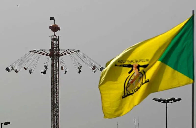 A flag of Iraq's pro-Iran paramilitary group Hezbollah Brigades (Kataib Hezbollah), flutters against the backdrop of people taking a swing ride at an amusement park during Eid al-Fitr celebrations, marking the end of the Muslim holy fasting month of Ramadan, in Baghdad on March 21, 2026. (credit: AHMAD AL-RUBAYE / AFP via Getty Images)