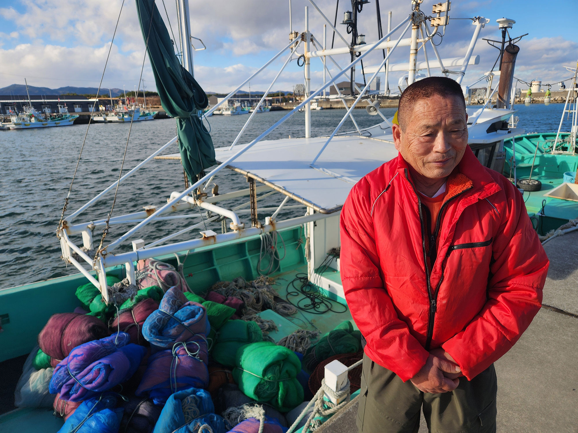 A grim faced Japanese man in a red jacket, perhaps 60, stands on a dock near his fishing boat on a sunny day 