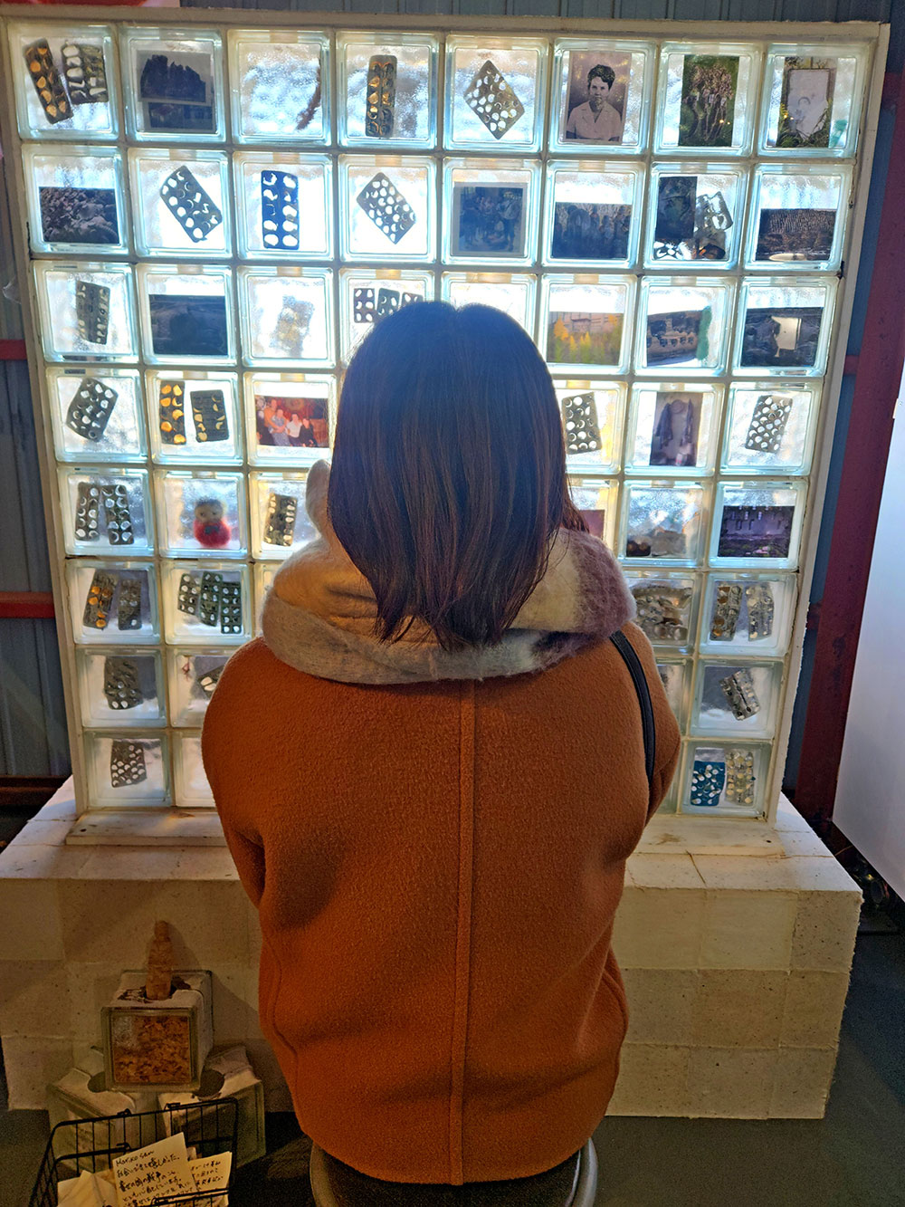 A Japanese woman in a red jacket faces away from the camera as she looks at a square containing many smaller squares, each containing a small artifact.