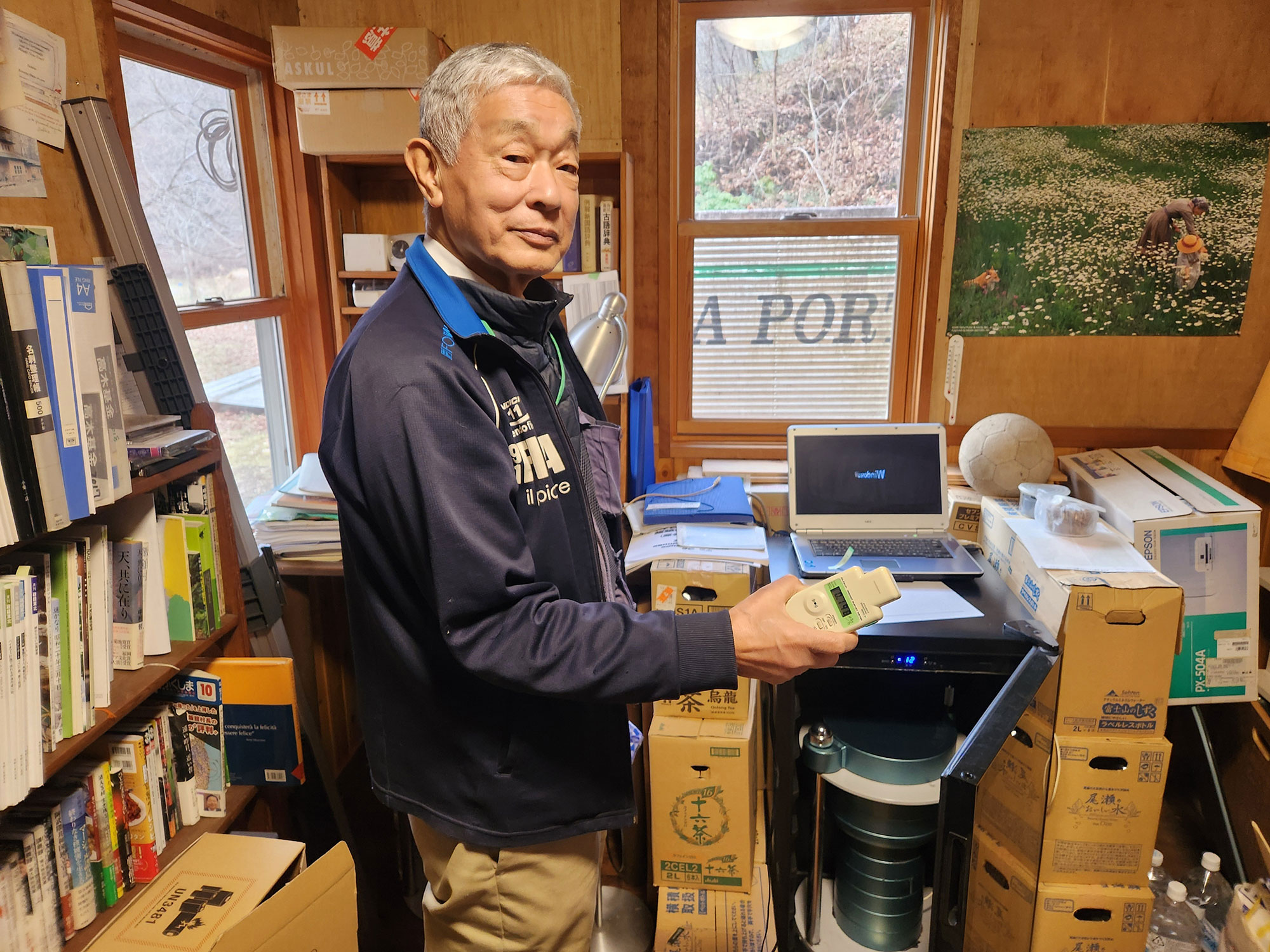 A sixty-ish Japanese man stands in a room cluttered with boxes, books, and files and wears a bemused expression.