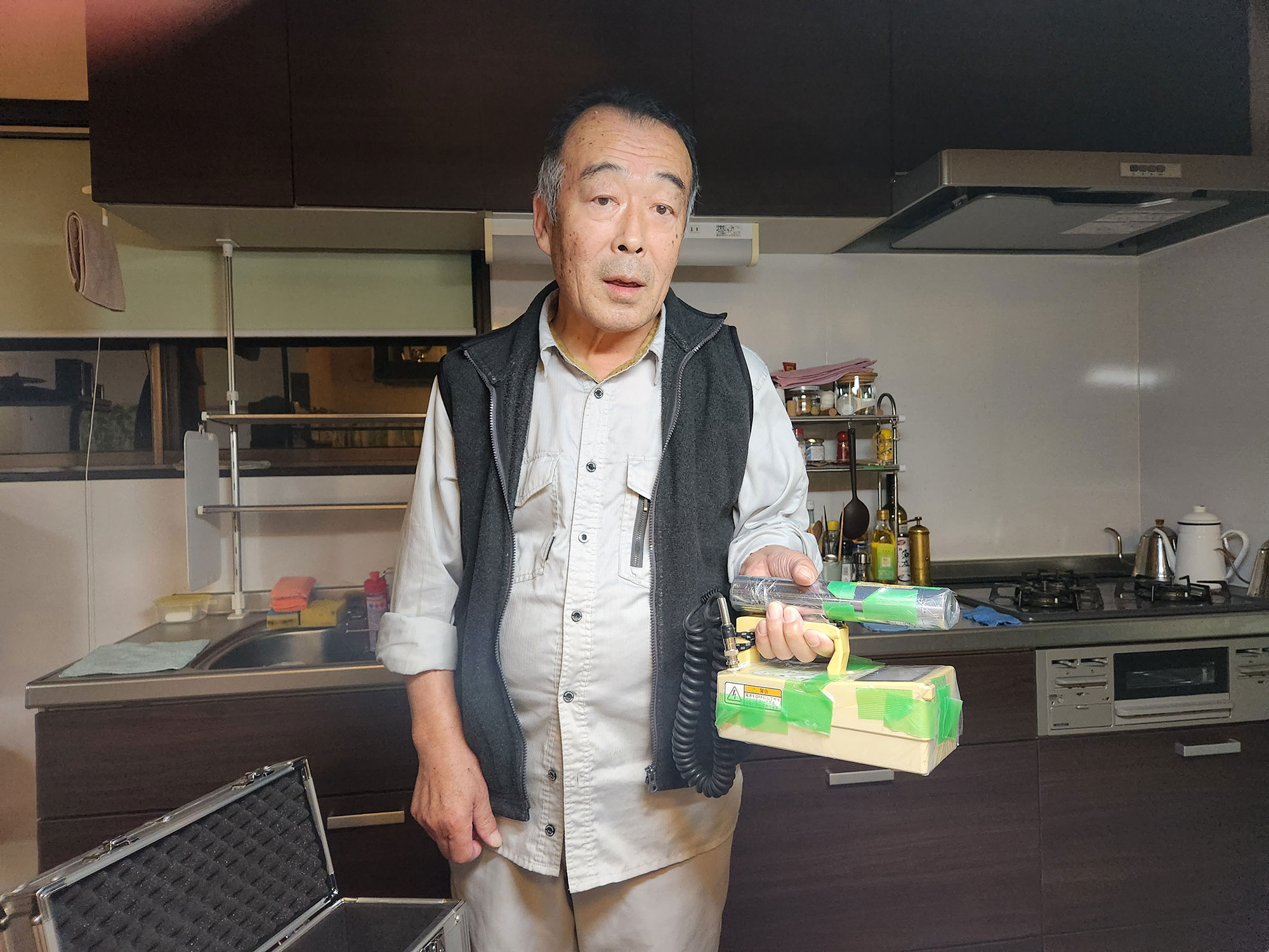A balding Japanese man stands in his kitchen holding a taped up geiger counter