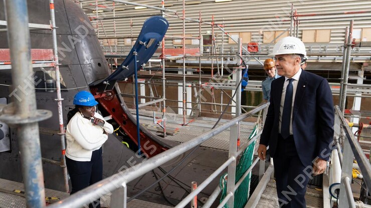 Mark Carney, wearing a hard hat, looks at a submarine being built as an employee looks at him while touring a facility.