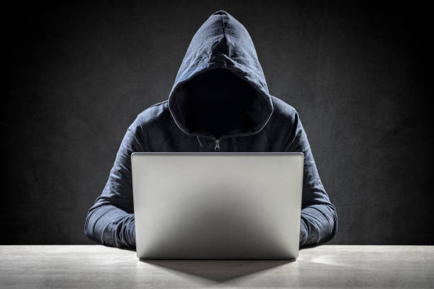 Illustration of a hooded figure using a laptop in a dark setting, representing hacking or cybercrime.
