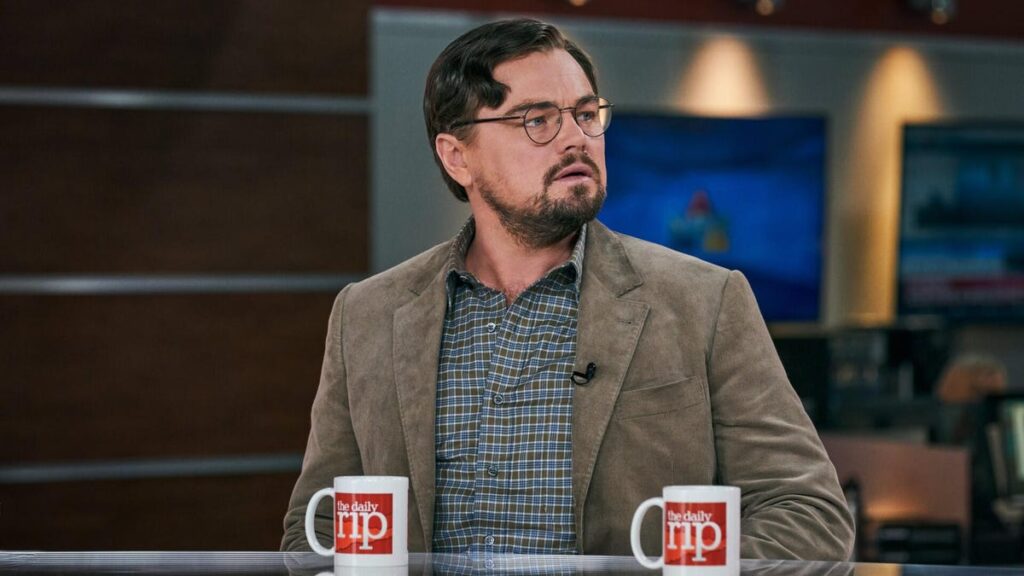 Production still from Don't Look Up of Leonardo DiCaprio sitting at a news desk in a schlubby suit and glasses.