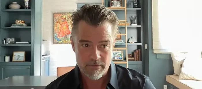 A man sits in a home office, looking towards the camera, with shelves and artwork in the background