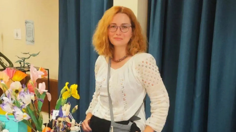 A woman with light red hair smiles at the camera. She is wearing a white shirt and standing behind a table covered in flowers.