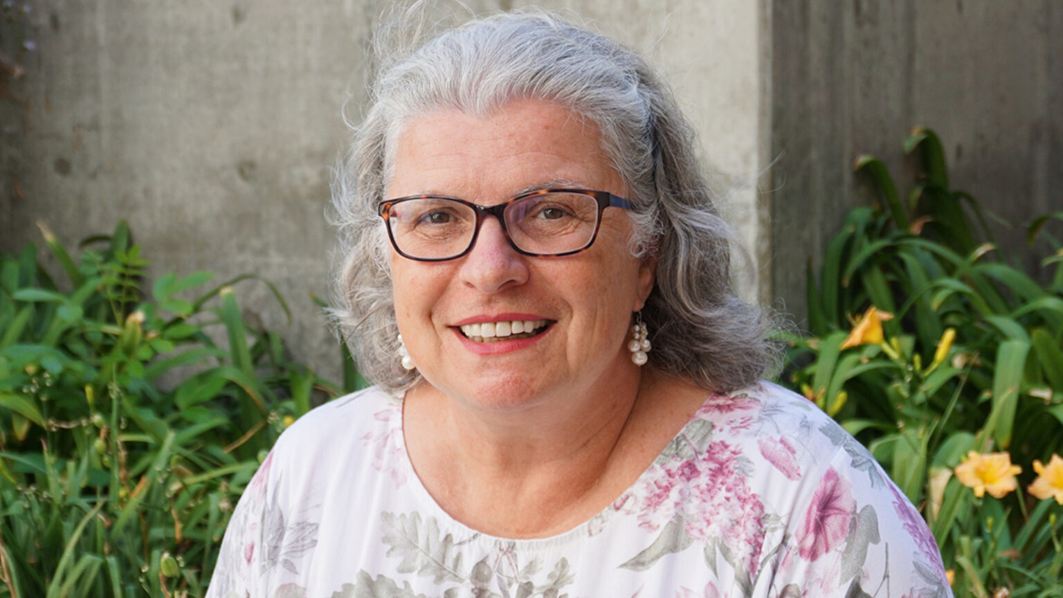 A portrait of a smiling woman in a floral print dress with some greenery and flowers in the background.