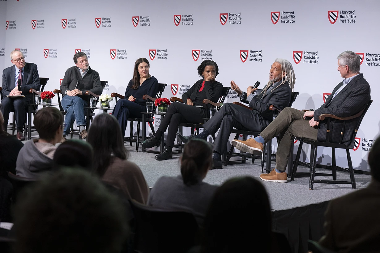 Six people sitting in chairs, one with a microphone speaking to a crowd, in front of a backdrop with the Harvard Radcliffe Institute logo on it