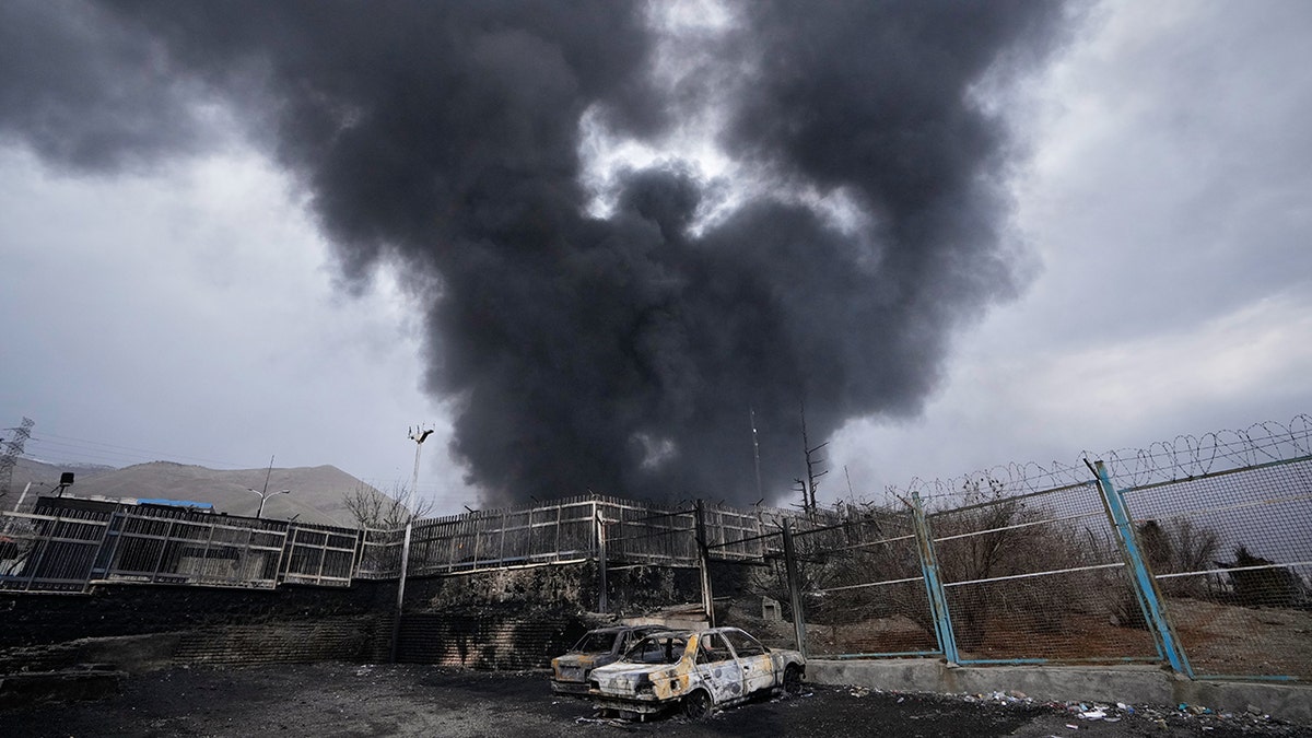 A thick plume of smoke rising from an oil storage facility into a cloudy sky.