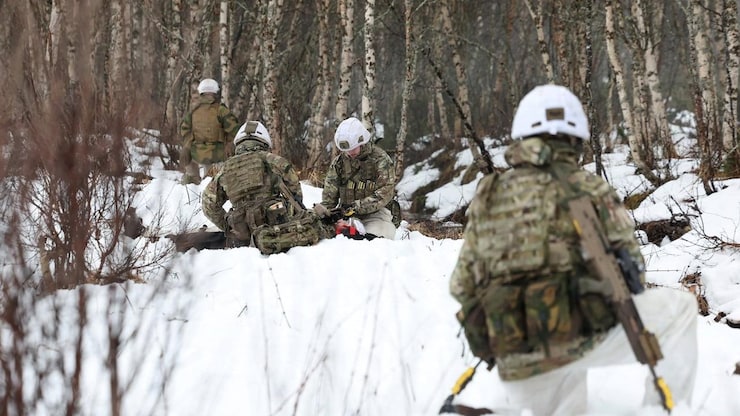 Soldiers in military attire preparing their gear in the snow near the woods.