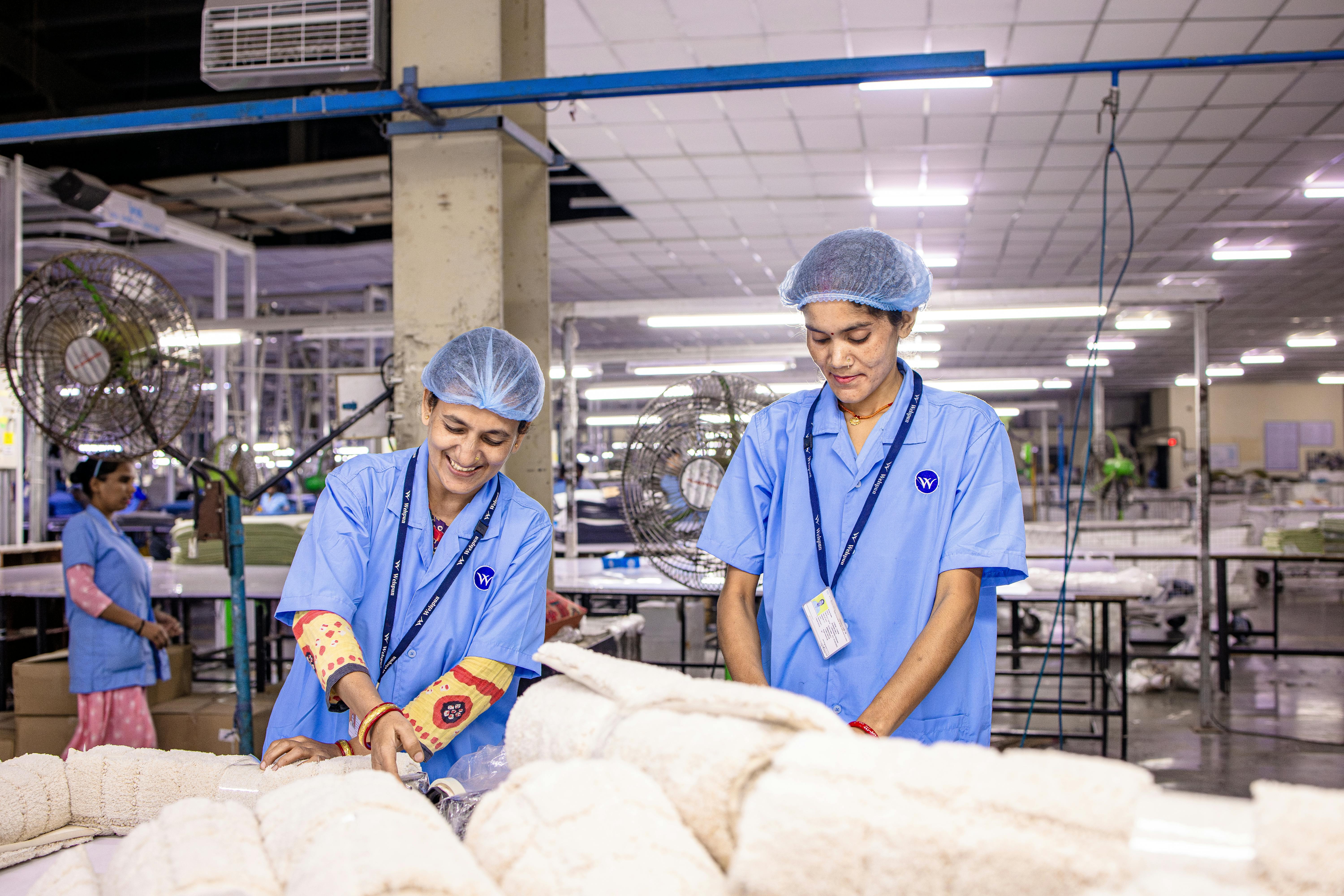 Factory workers in blue uniforms and hairnets fold textiles on a production floor.