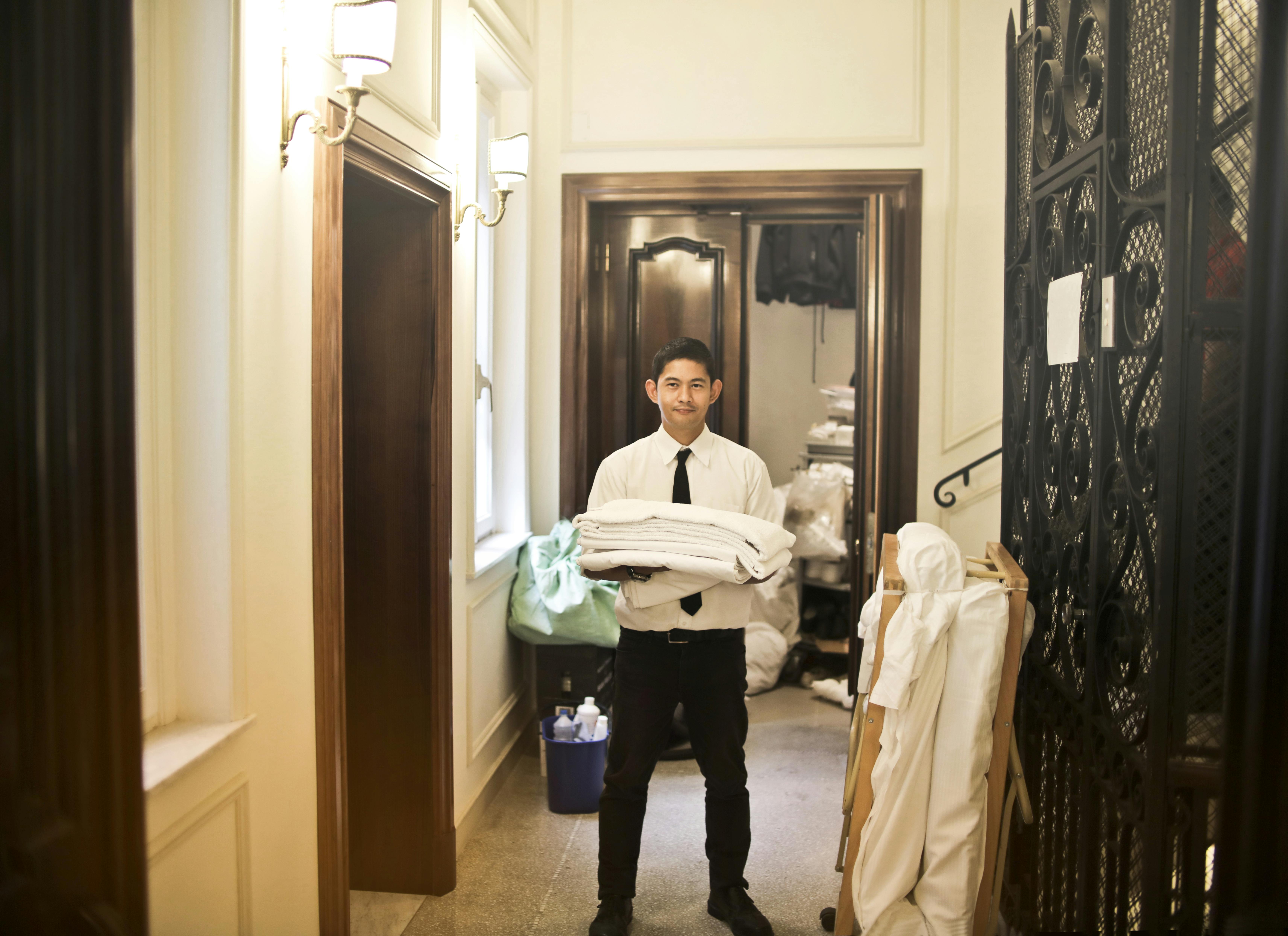 Hotel staff member carrying folded towels stands in a hallway near a laundry room.