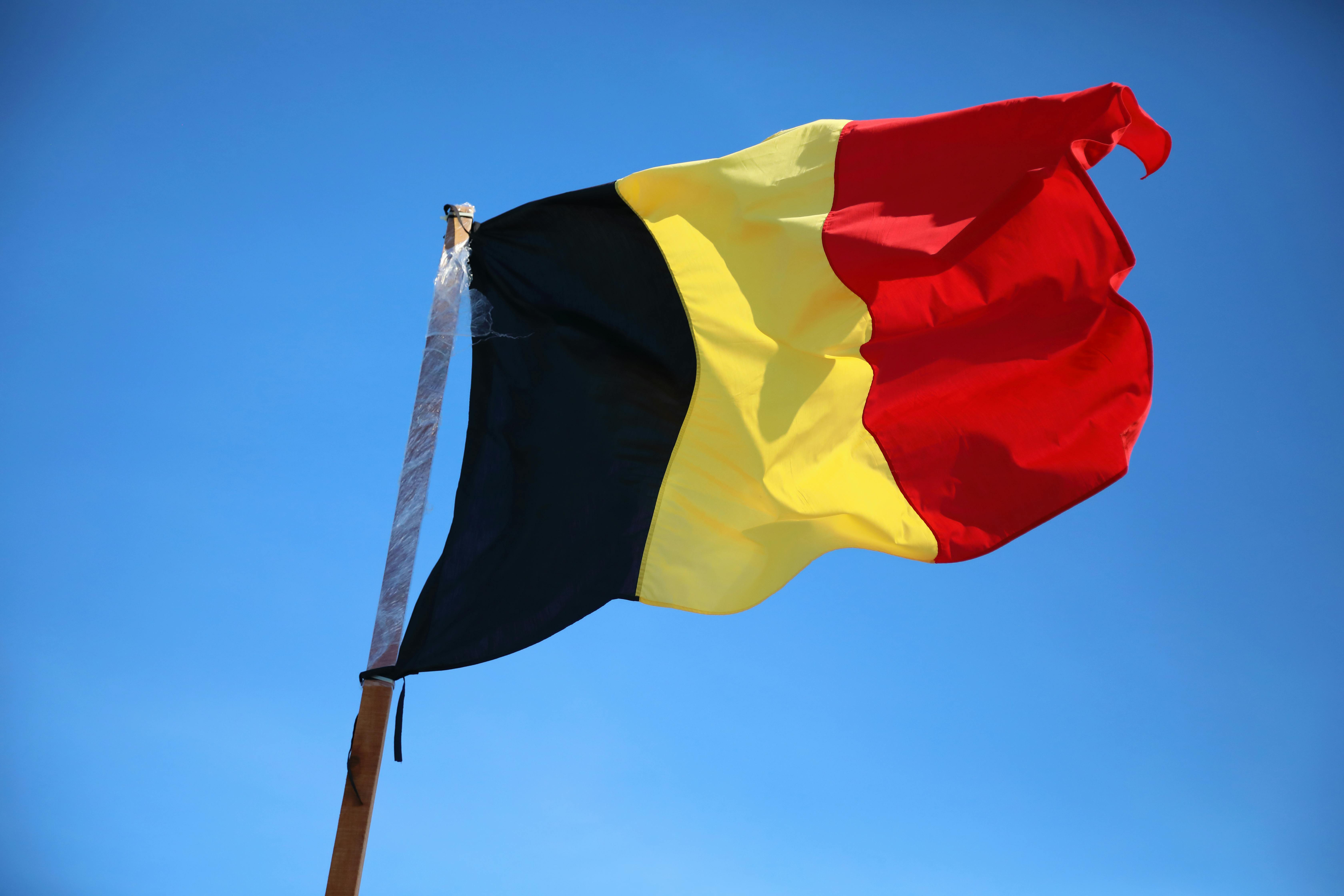Belgium national flag with black, yellow, and red stripes waving on a flagpole against a clear blue sky.