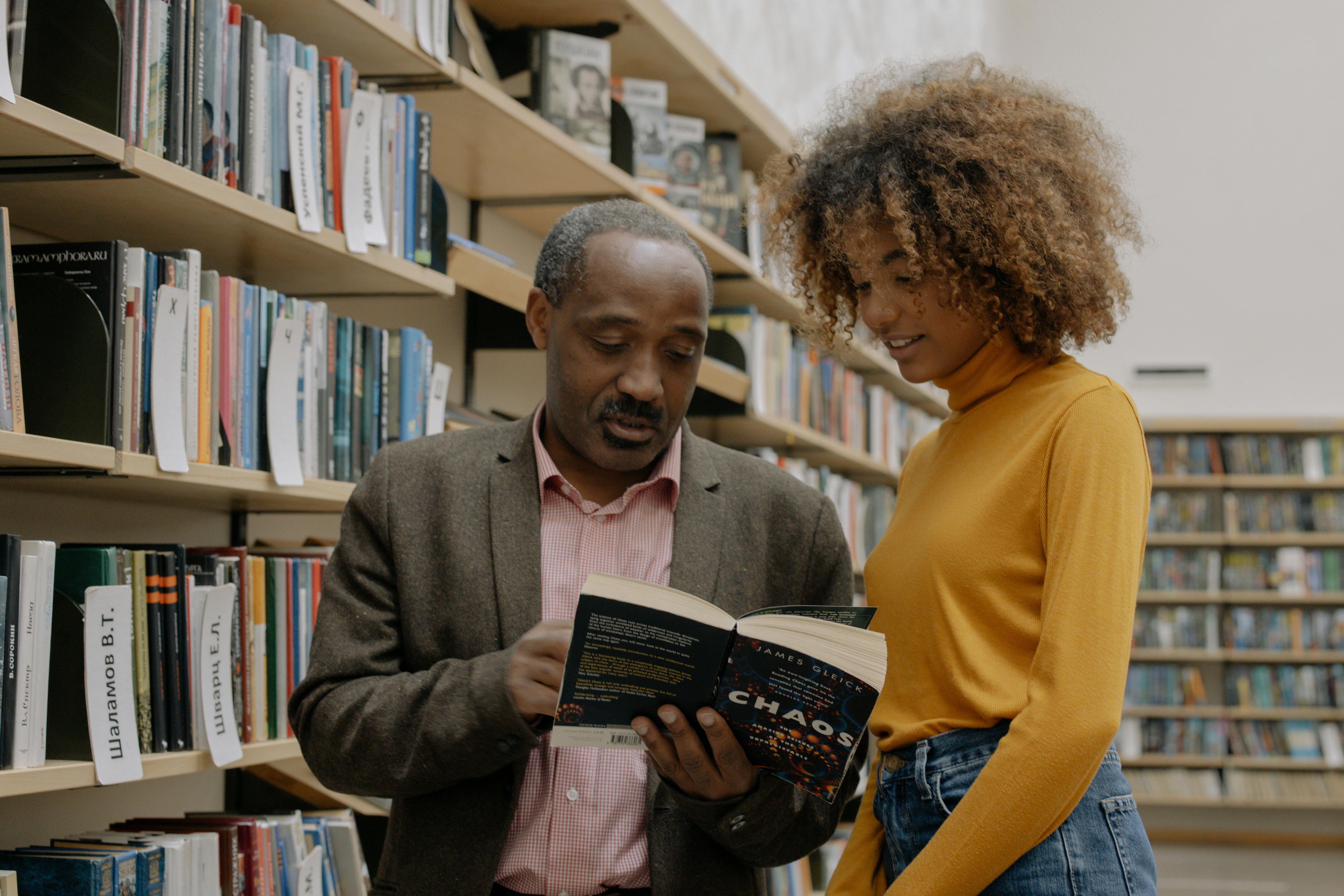 Man and young woman reading a book together beside shelves in a library.
