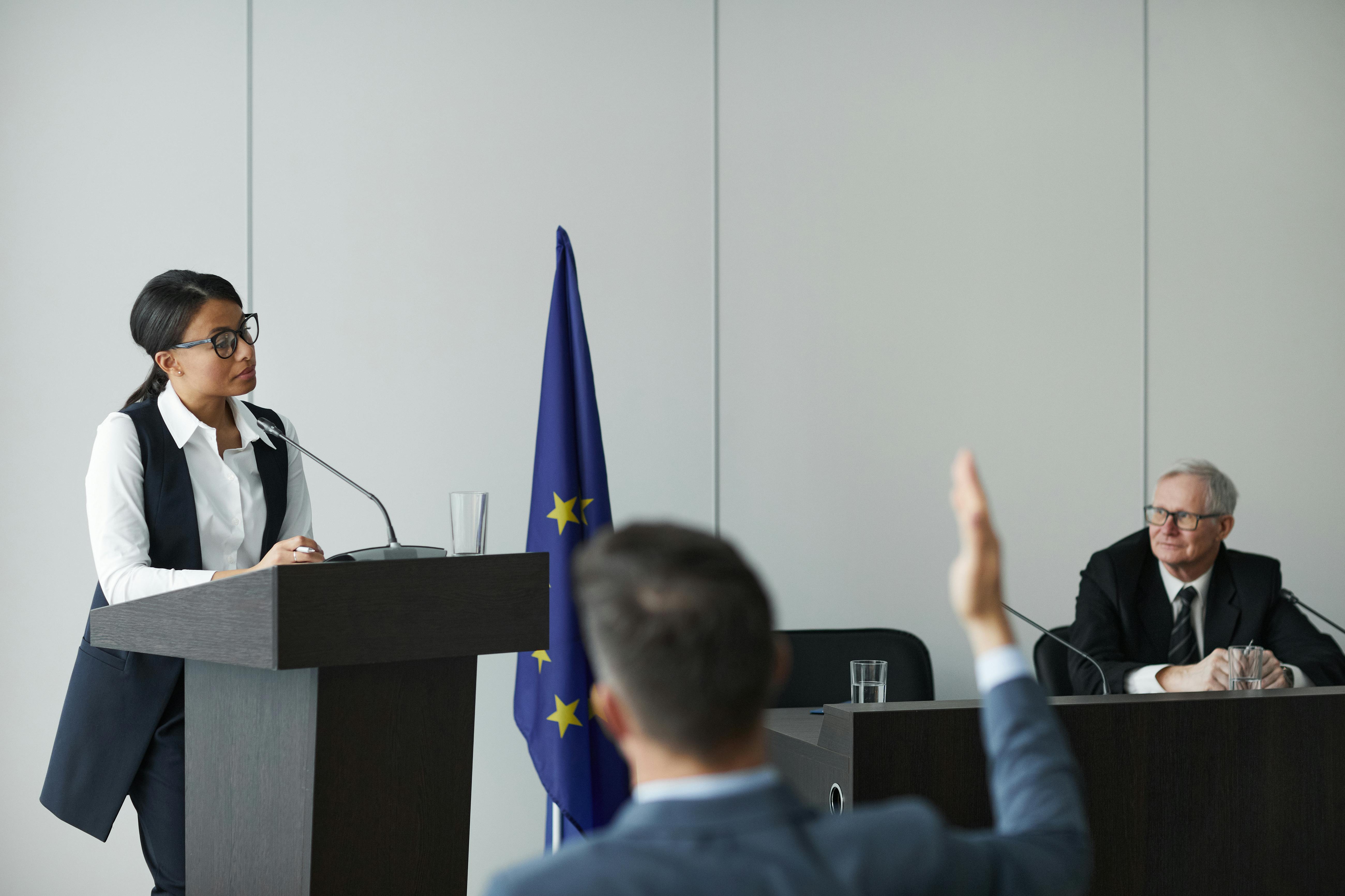 Official speaking at a podium during a press conference with a European Union flag in the background.
